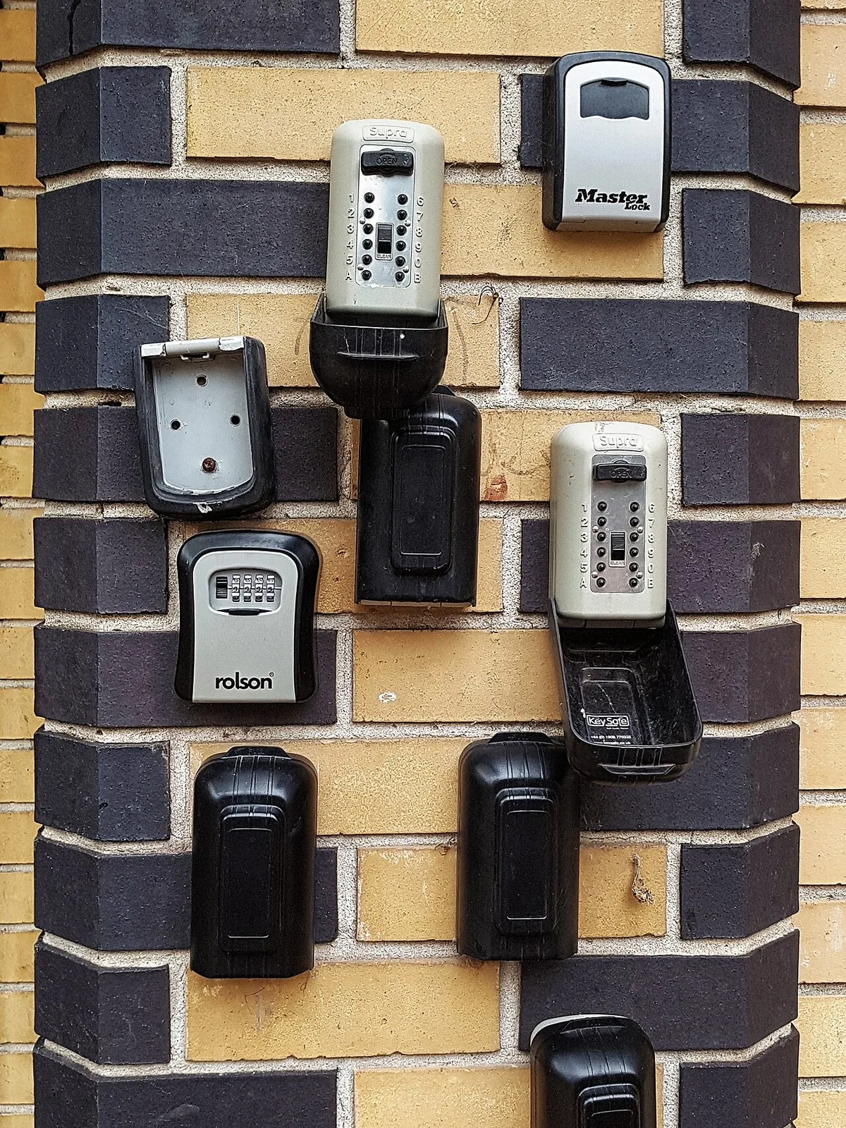 Vijf sleutelkluisjes met cijfertoetsen naast elkaar bevestigd op een muur van gele en donkere baksteen.