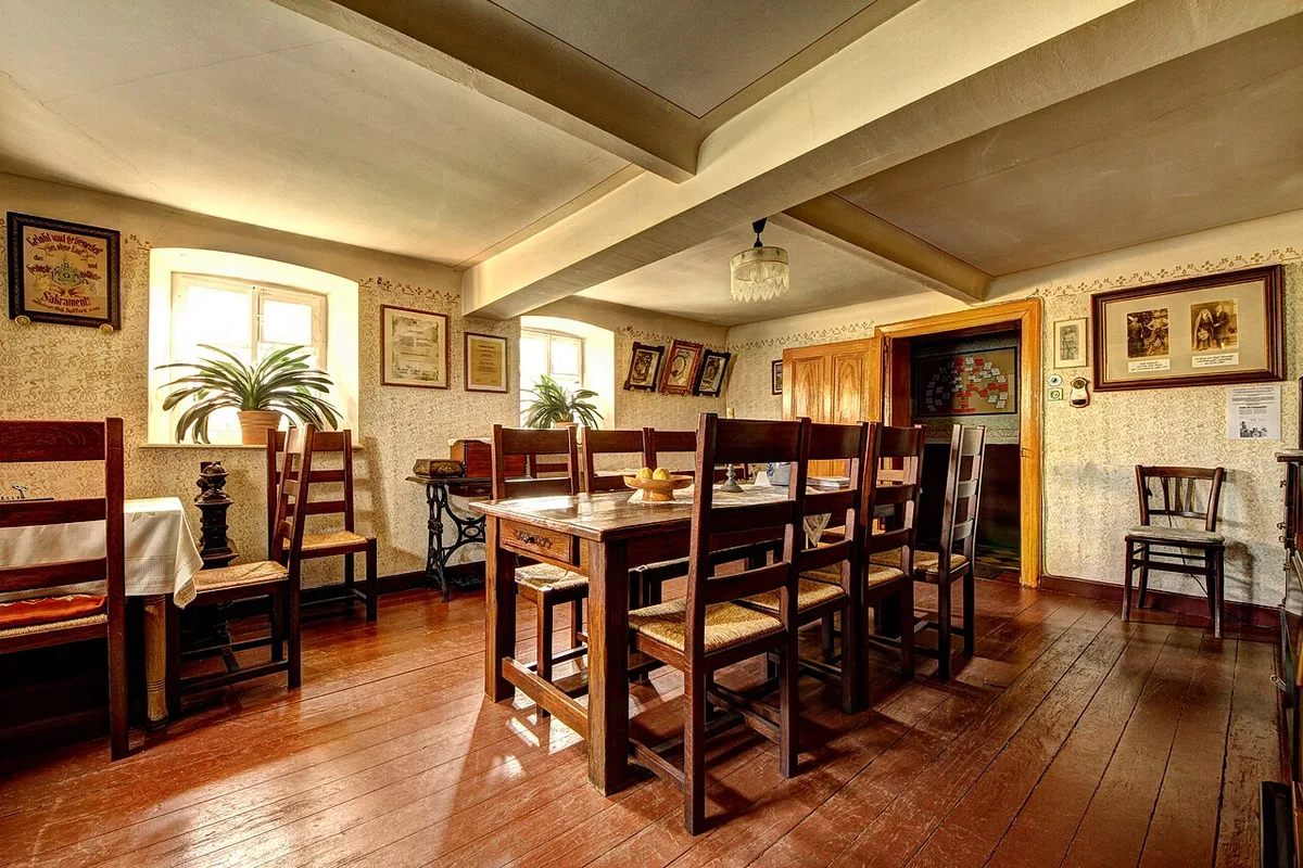 Een landelijke woonkamer met donkere houten balken aan het plafond, een lange eettafel met stoelen en houten lambrisering langs de wanden.